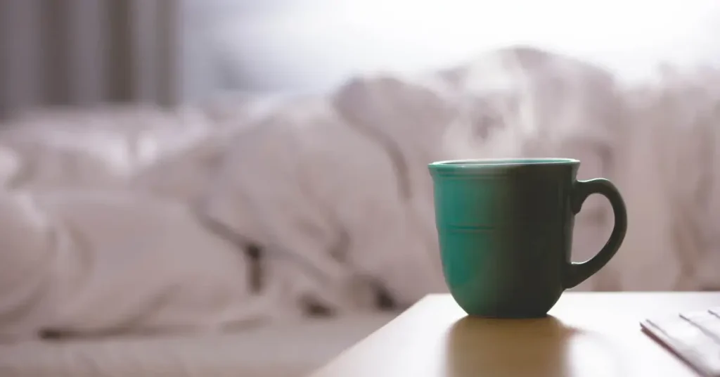 A picture of a cup in front of an undone bed.