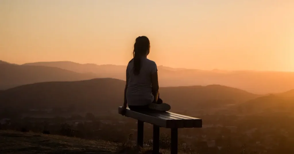 A woman is sitting on a bench and enjoying the view of hills with the morning sun.